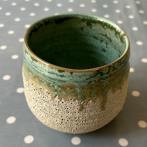 Puede incluir: Una taza de cerámica con un exterior marrón claro moteado y un glaseado verde en el interior y el borde. El glaseado tiene un aspecto jaspeado y desigual.