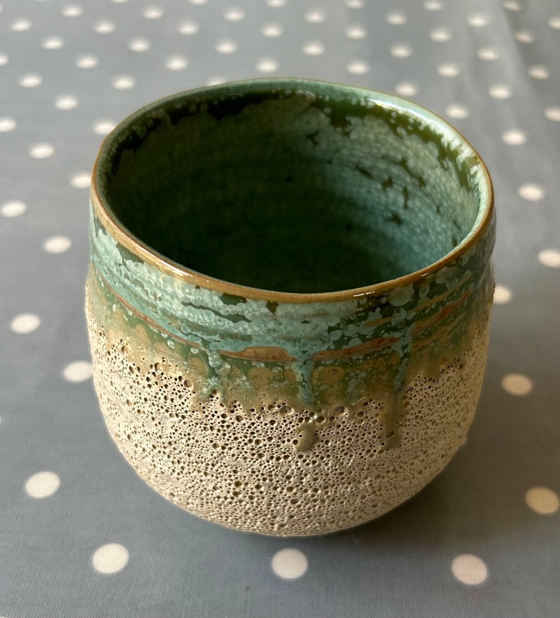 May include: A ceramic mug with a speckled, light brown exterior and a green glaze on the inside and rim. The glaze has a mottled, uneven appearance.