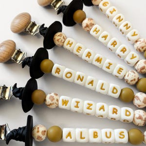 May include: Close-up of several baby pacifier clips. Each clip features a black witch hat, wooden clip, and a series of white square beads with gold lettering spelling out names like "Harry" and "Hermie". The clips also have decorative beads.