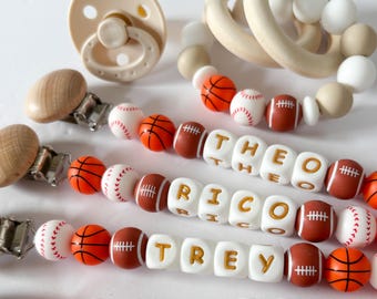 May include: A selection of baby items, including a dummy, wooden teething rings, and personalised dummy clips. The clips feature beads shaped like baseballs, basketballs, and American footballs, with names spelled out in lettered blocks. The colour scheme includes beige, white, and brown.