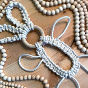 May include: A white macrame wall hanging with a wooden ring and wooden beads. The macrame is made with a knotting technique and has a delicate, airy feel.