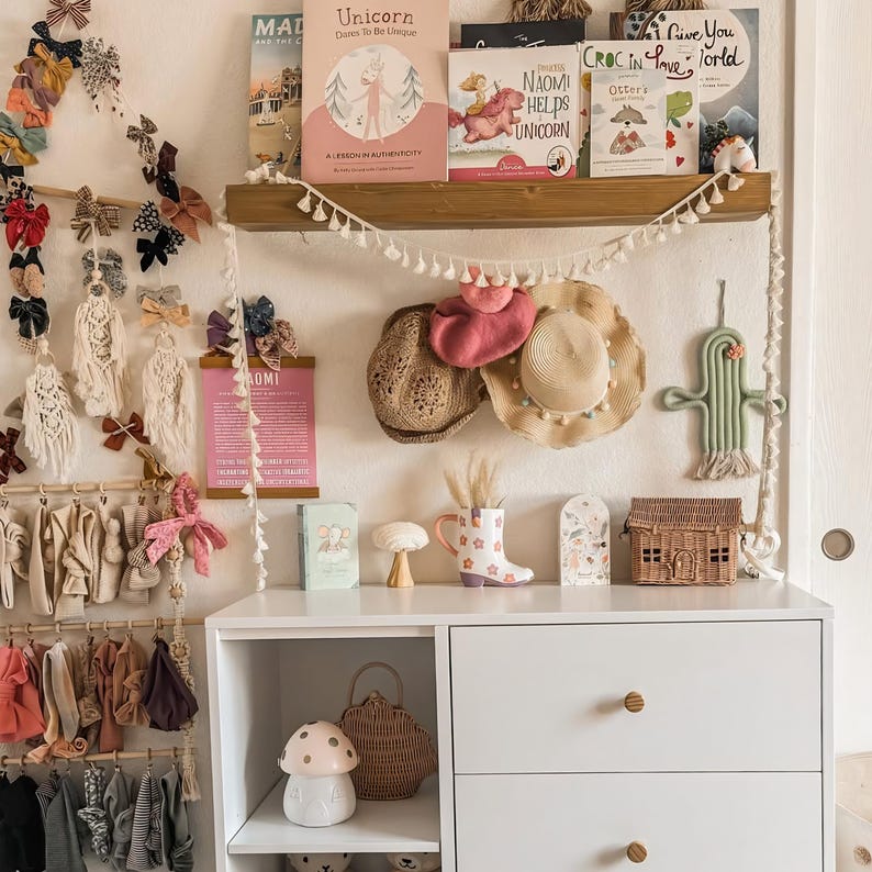 May include: A white dresser with wooden knobs displays children's accessories and decor. A shelf above holds books, hats, and a cactus-shaped wall hanging. Bows and clothing hang on the wall, creating a whimsical nursery or children's room.
