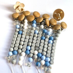 May include: A set of 10 silicone baby teethers with wooden clips. Each teether is made with gray, blue, and white beads and features a personalized name or the word "COWBOYS".
