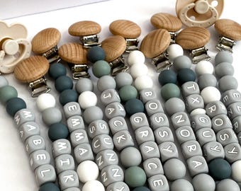 Op de afbeelding: Een collectie speenkoorden met ronde siliconen kralen in saliegroen, grijs en wit. Elke clip heeft een ronde houten sluiting en een metalen klem. De kralen zijn gepersonaliseerd met namen zoals MABEL, MIKE, EMMA, SUNNY, JACK, GRACE, NORA, GUY en ROSY.