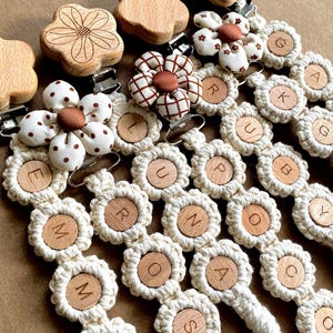 May include: A set of five crocheted baby teethers with wooden flower accents. Each teether features a different letter of the alphabet, including E, M, R, O, and A. The teethers are made with a cream-colored yarn and have a brown and white polka dot flower accent.