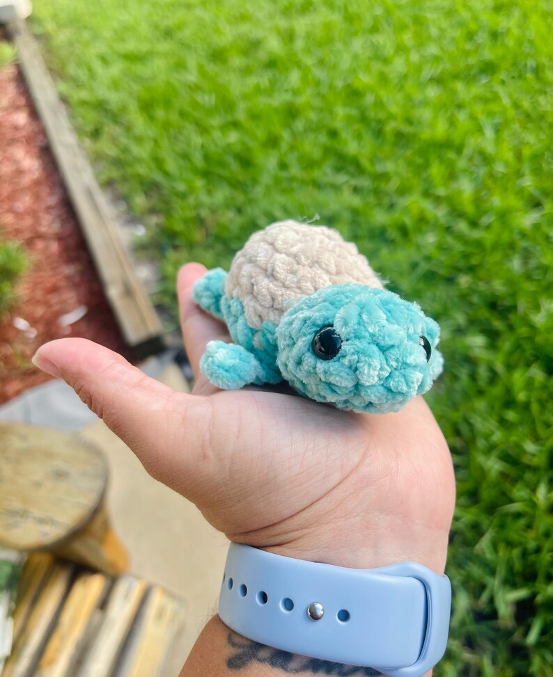 May include: A small crocheted turtle toy with a light blue shell and a beige head. The turtle is sitting in the palm of a hand.