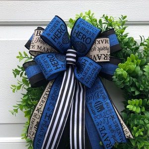 May include: A blue and white wreath with a blue ribbon bow. The ribbon has the words "Protect & Serve" and "Hero" printed on it. The wreath is decorated with green leaves.