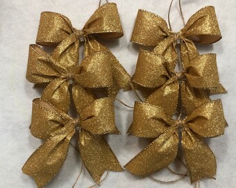 May include: Six gold glitter bows with twine attached. The bows are arranged in a stack, with the top bow facing the camera.