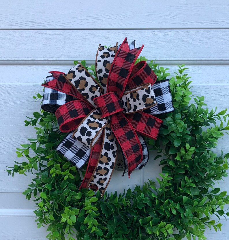 May include: A green wreath with a large bow made of red and black buffalo plaid ribbon, black and white checkered ribbon, and brown and tan leopard print ribbon.