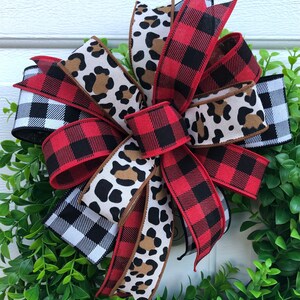 May include: A red and black buffalo plaid ribbon, a black and white gingham ribbon, and a brown and white leopard print ribbon are tied together in a bow. The bow is on a green wreath.