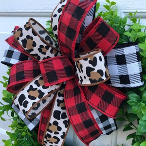May include: A red and black buffalo plaid ribbon, a black and white buffalo plaid ribbon, and a brown and white leopard print ribbon are tied together in a bow. The bow is on a green leafy wreath.