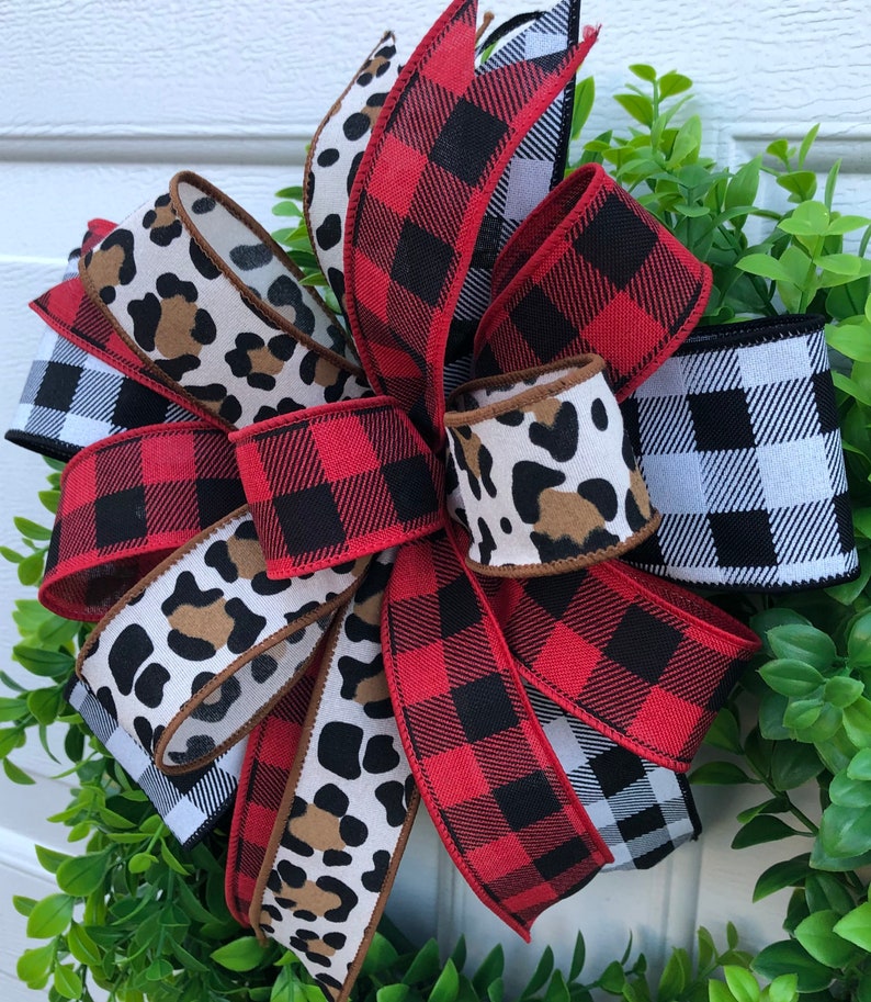 May include: A red and black buffalo plaid ribbon, a black and white buffalo plaid ribbon, and a brown and white leopard print ribbon are tied together in a bow. The bow is on a green leafy wreath.