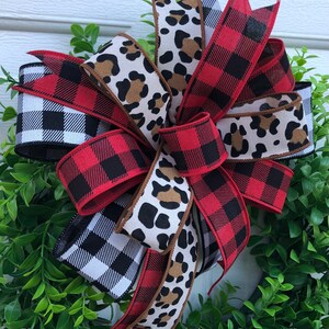May include: A red and black buffalo plaid ribbon, a black and white gingham ribbon, and a brown and white leopard print ribbon are tied together in a bow. The bow is on a green wreath.