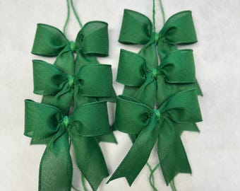 May include: Six green fabric bows with attached green strings. The bows are made of a sheer, slightly shiny material. The bows are tied in the center and have a classic bow shape. The background is a plain white surface.
