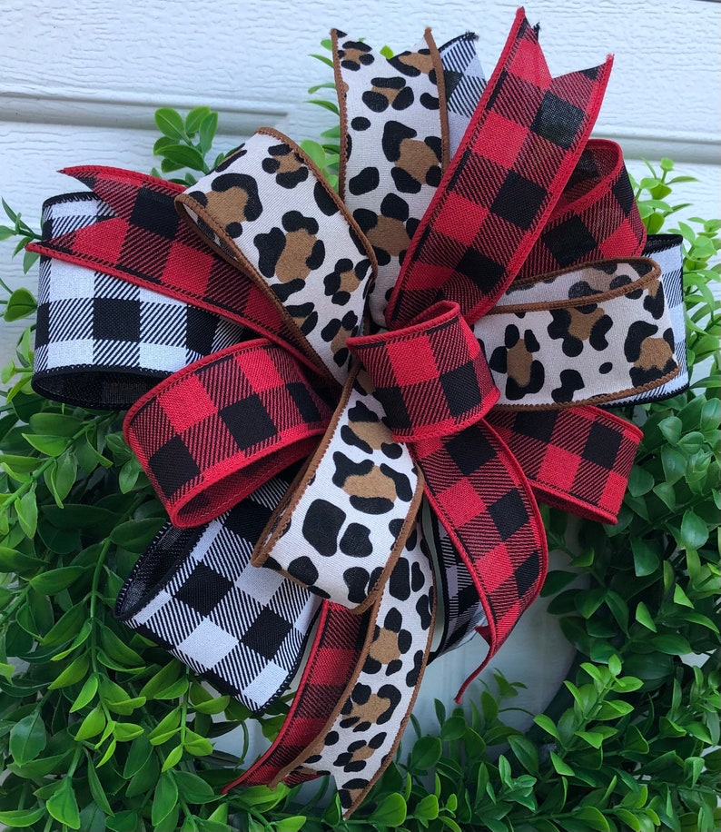 May include: A red and black buffalo plaid ribbon, a black and white gingham ribbon, and a brown and white leopard print ribbon are tied together in a bow. The bow is on a green wreath.