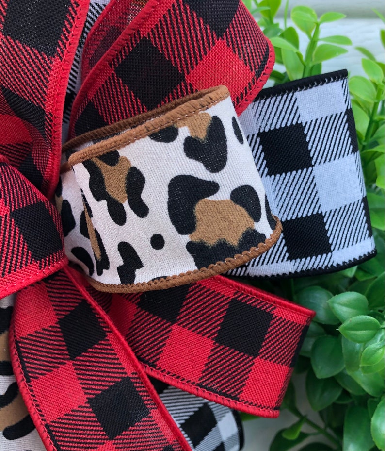 May include: A close-up of a red and black buffalo plaid ribbon, a white and black buffalo plaid ribbon, and a brown and white leopard print ribbon. The ribbons are tied together in a bow.