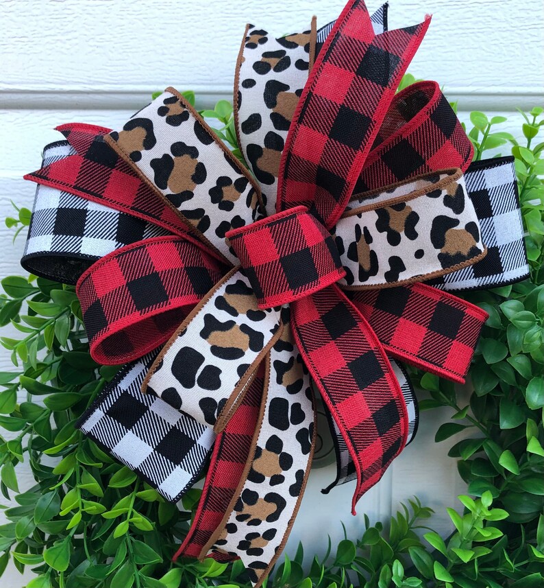 May include: A red and black buffalo plaid ribbon, a black and white gingham ribbon, and a brown and white leopard print ribbon are tied together in a bow. The bow is on a green wreath.