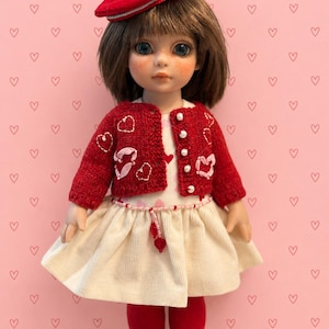 May include: A doll with brown hair wearing a red beret with a white pom-pom, a red knit cardigan featuring embroidered hearts and white buttons, and a cream-colored dress with a beaded waist detail. The doll also wears red tights and red shoes.