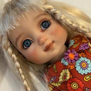 May include: A doll with long blonde hair styled in braids, featuring bright blue eyes, long eyelashes, and light freckles on the cheeks. The doll wears a rust-colored fabric garment with a colorful floral pattern and a small purple flower detail.