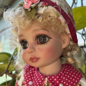 May include: A doll with curly blonde hair, large dark eyes, and long eyelashes. The doll wears a floral-patterned headpiece with a pink fabric flower and a pink-and-white polka-dot collar with small white buttons on a floral-patterned garment.