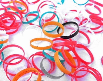 May include: A collection of colorful plastic rings in various sizes and colors, including red, orange, yellow, green, blue, white, and black. The rings are arranged in a random pattern on a white surface.