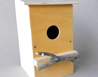 May include: A wooden birdhouse with a white roof and a round entrance hole. A small branch is attached to the front of the house.