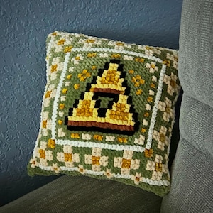 Peut inclure: Un coussin vert et jaune avec une image pixélisée d'une pyramide sur le devant. Le coussin a une bordure blanche et un motif à carreaux.