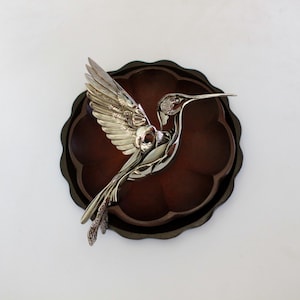 Puede incluir: Una escultura de colibrí de metal plateado con detalles intrincados en relieve en plumas y alas, colocada sobre un cuenco de madera circular de color marrón oscuro con borde festoneado, acabado mate liso y un interior profundo y redondeado.