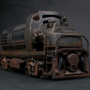Pode incluir: Modelo detalhado de uma locomotiva a diesel vintage, com uma aparência desgastada e enferrujada. O comboio castanho escuro e preto tem parafusos, tubos e um corrimão frontal visíveis. O modelo está sobre um fundo escuro.