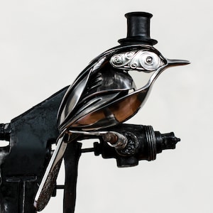 Peut inclure: Une sculpture métallique fantaisiste représentant un oiseau coiffé d'un chapeau haut de forme. L'oiseau est fabriqué à partir de couverts, avec un design détaillé. La sculpture est montée sur une base en métal noir, créant une œuvre d'art unique.