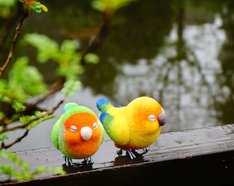 May include: Two small felt bird figurines, one green, orange, and blue, the other yellow, orange, and blue, perched on a wooden surface.