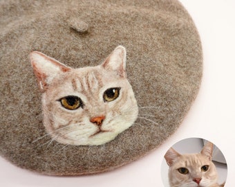 May include: A brown felt beret with a felted cat face design. The cat has light brown fur with darker stripes and yellow eyes.