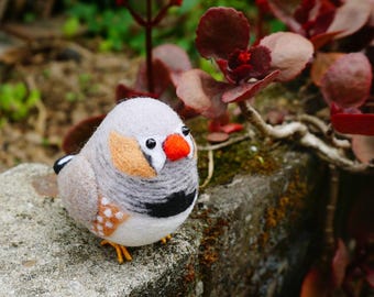May include: A needle-felted zebra finch figurine featuring a round, light gray body, a bright orange beak, black eye beads, and orange cheek patches. The bird has a white chest with a black horizontal band and white-spotted brown wing feathers.