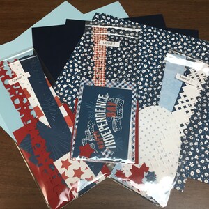 Puede incluir: Un conjunto de tres diseños de papel para álbum de recortes con diseños patrióticos rojo, blanco y azul. Los diseños incluyen una tarjeta con el texto "Feliz Día de la Independencia", un papel en forma de corazón con lunares blancos y una variedad de papeles estampados rojo, blanco y azul.