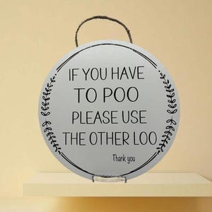 May include: A round white sign with black text and a black floral border. The sign reads "If you have to poo please use the other loo. Thank you."