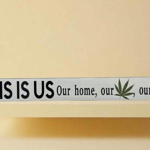 May include: A rectangular wooden sign with the text "THIS IS US" in black letters. The sign also includes the phrases "Our home, our, our life." and a green cannabis leaf. The sign is on a light-coloured shelf.