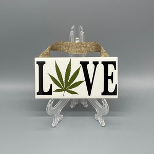 May include: A rectangular white ceramic sign with the word "LOVE" in black capital letters. A green cannabis leaf is in place of the "O". A tan burlap ribbon is attached for hanging.
