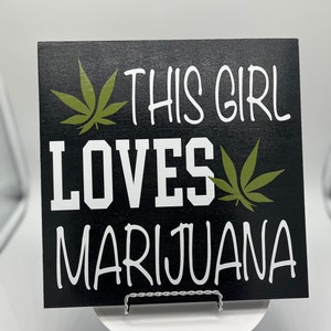 May include: Black wooden sign with white text that reads "This girl loves marijuana" with green marijuana leaf graphics.
