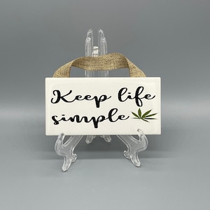 May include: White ceramic sign with the text "Keep life simple" and a green marijuana leaf. The sign is hanging from a brown twine rope.