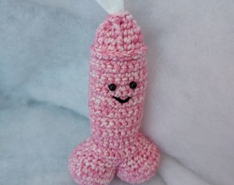 May include: A pink and white crocheted tissue holder shaped like a penis, with a smiling face and a white tissue emerging from the top. The handmade item is set against a soft, white background.