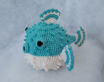 May include: A crocheted pufferfish toy with a blue and white striped pattern. The toy has a white belly and black eyes.