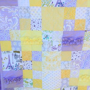 May include: A patchwork quilt with a yellow and purple color scheme. The quilt is made of various fabrics with different patterns, including floral, polka dot, and geometric designs. The quilt has a ruffled edge and features a small Eiffel Tower design.
