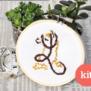May include: Embroidery hoop kit featuring a brown and yellow llama design. The kit includes all the materials needed to complete the project. The word "kit" is printed in white on a red circle.