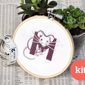 May include: A purple and white embroidery kit featuring a mouse in the shape of the letter M. The kit is displayed in a wooden embroidery hoop.