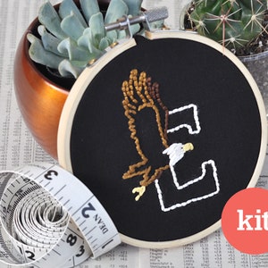 May include: A DIY embroidery kit featuring a black embroidery hoop with a white outline of the letter "E" and a brown and white embroidered eagle inside. The kit includes a white measuring tape and the word "kit" in red.