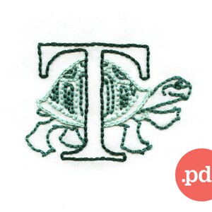 以下が含まれることがあります： 緑の亀のデザインが施された刺繍された文字T。亀は黒い糸で縁取られており、緑と白の甲羅をしています。