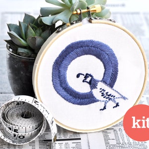 Puede incluir: Un bastidor de bordado con una letra "Q" bordada en azul y un diseño de pájaro. La palabra "kit" está en un círculo rojo. Una planta suculenta en una maceta oscura y una cinta métrica están en el fondo.