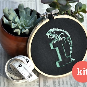 May include: A black embroidery hoop with a green iguana design. The hoop is 8 inches in diameter and is surrounded by a measuring tape. The word "kit" is written in red on a white background.
