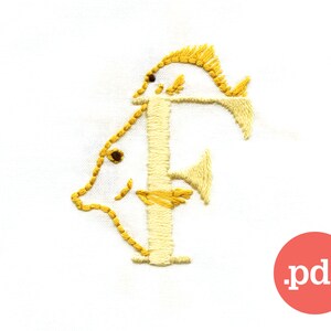 May include: A yellow embroidered letter F with two fish designs. The letter is stitched on a white fabric background.