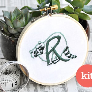 May include: An embroidery kit featuring the letter "R" with a bear design. The kit includes a hoop, fabric, and embroidery floss. The word "kit" is printed in red on a white circle.